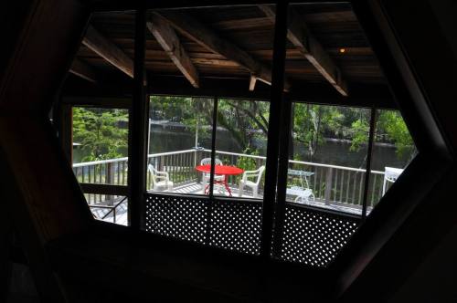 View from a hexagonal window overlooking a deck with a red table by a serene river surrounded by greenery.