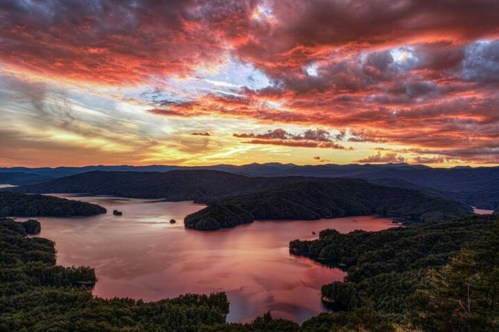 A stunning sunset over a tranquil lake, surrounded by lush mountains and vibrant clouds in the sky.