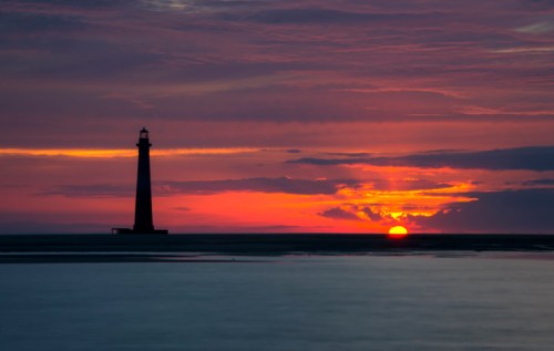 A silhouette of a lighthouse against a vibrant sunset with colorful clouds and a calm sea.