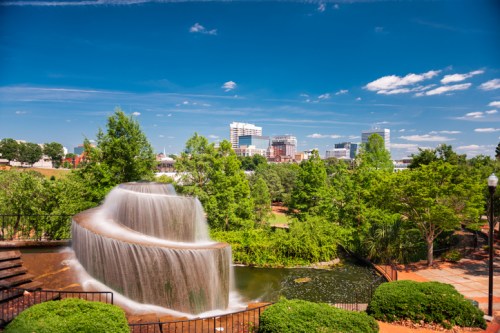 A cascading waterfall flows into a serene pond, surrounded by lush greenery and a city skyline in the background.