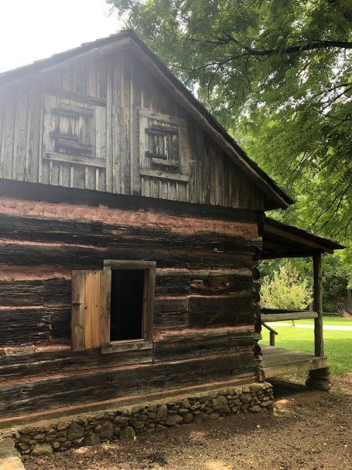 A rustic wooden cabin with a sloped roof, featuring a small window and a porch, surrounded by greenery.