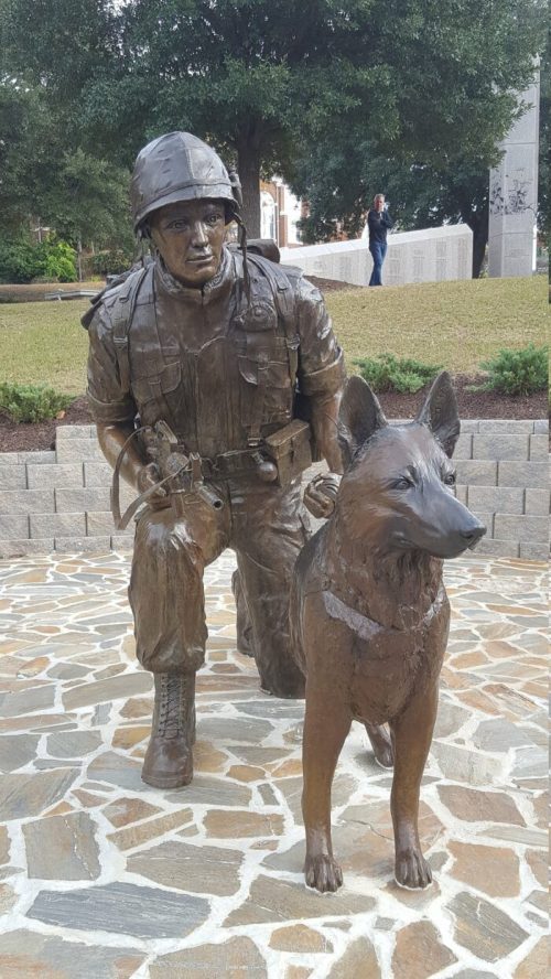 A bronze statue of a soldier kneeling beside a military dog, set in a landscaped area with trees in the background.