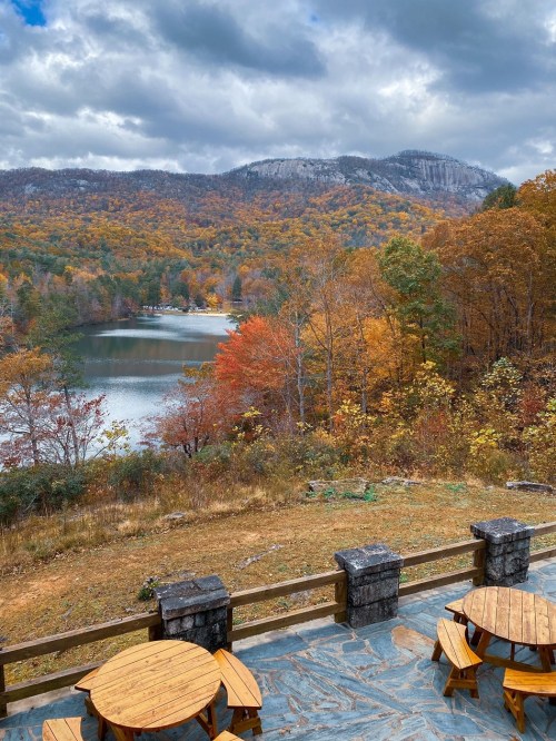 Scenic view of a lake surrounded by autumn foliage and mountains under a cloudy sky, with wooden tables in the foreground.