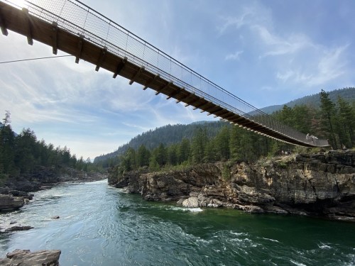 A suspension bridge spans a river, surrounded by rocky cliffs and lush green trees under a clear blue sky.