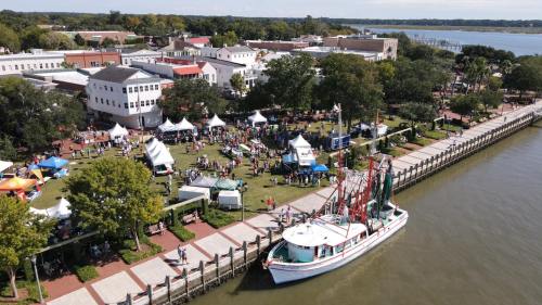 Aerial view of a waterfront festival with tents, people, and a boat docked along the riverbank.