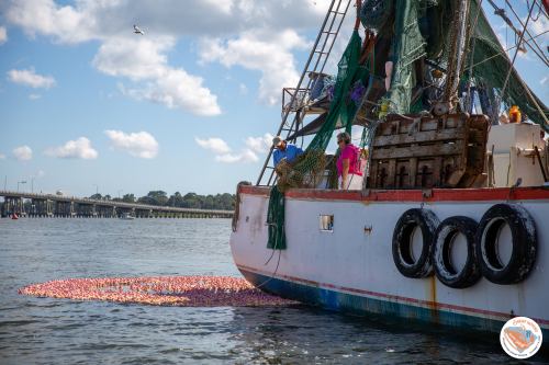 A fishing boat with nets on the side floats near a colorful cluster of objects in the water under a blue sky.