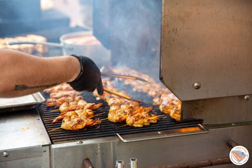A chef uses tongs to grill marinated chicken on a barbecue, with smoke rising in the background.