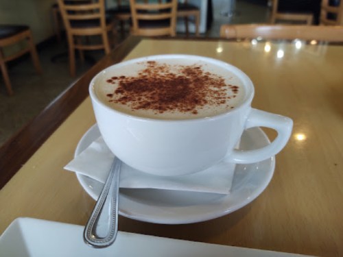 A white cup of cappuccino topped with cocoa powder, sitting on a saucer with a spoon beside it on a wooden table.