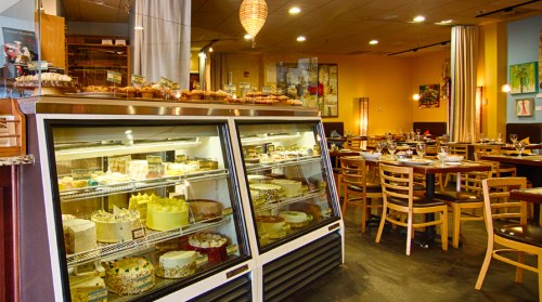A cozy restaurant interior featuring a display case of cakes and pastries, with tables set for dining.