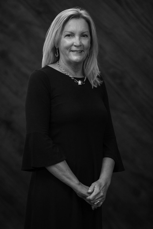 A woman in a black dress stands confidently, smiling, with her hands clasped in front of her against a textured background.