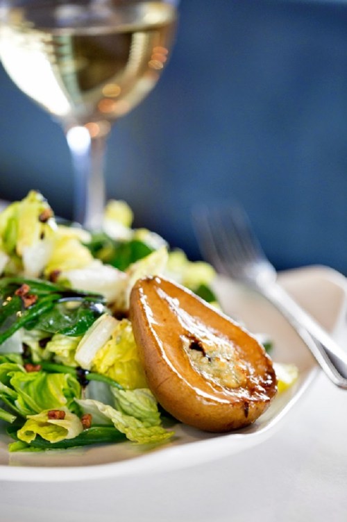 A plate of salad with greens and a grilled pear, accompanied by a glass of white wine in the background.