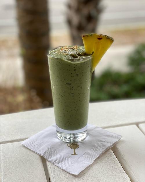 A tall glass of green smoothie topped with a pineapple slice, resting on a white napkin with a palm tree logo.