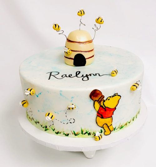 A whimsical cake featuring Winnie the Pooh, bees, and a honey pot, with the name "Raelynn" elegantly written on it.