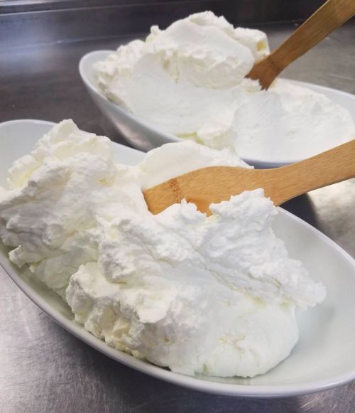 Two bowls of fluffy whipped cream with wooden spoons resting on top, set on a stainless steel surface.