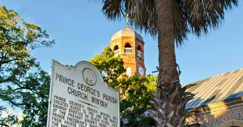 Historic sign for Prince George's Parish Church, Winyah, with a clock tower and palm tree in the background.