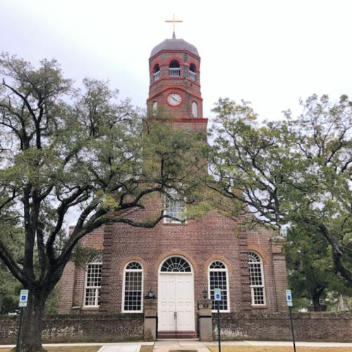 Historic brick church with a clock tower, surrounded by trees and a cloudy sky.