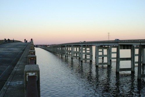 A long bridge stretches over calm water, reflecting the pastel colors of the sunset sky.