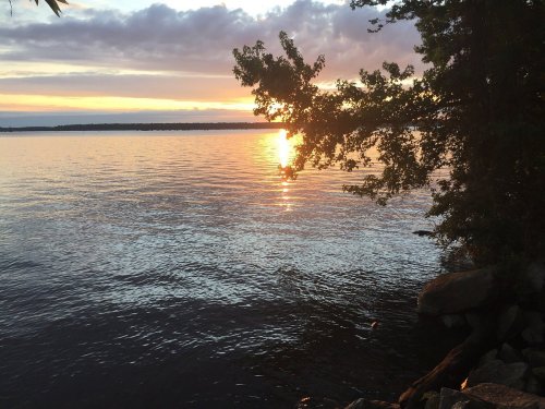 A serene sunset over a calm lake, with reflections on the water and trees framing the scene.