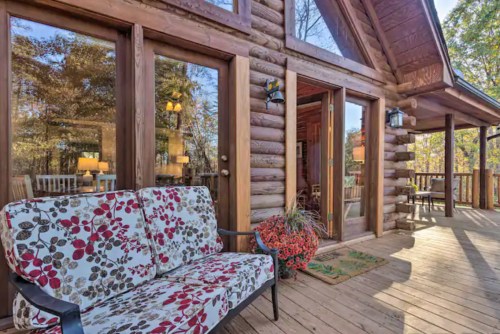 A cozy log cabin porch with a floral sofa, surrounded by trees and natural light streaming through large windows.