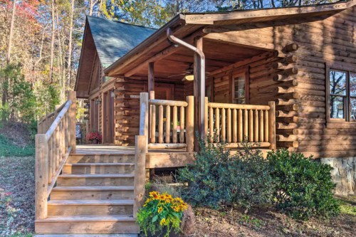 A rustic log cabin with a wooden porch and steps, surrounded by trees and colorful flowers.
