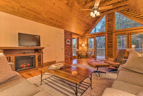 Cozy cabin living room with wooden walls, a fireplace, TV, and large windows overlooking a forested area.
