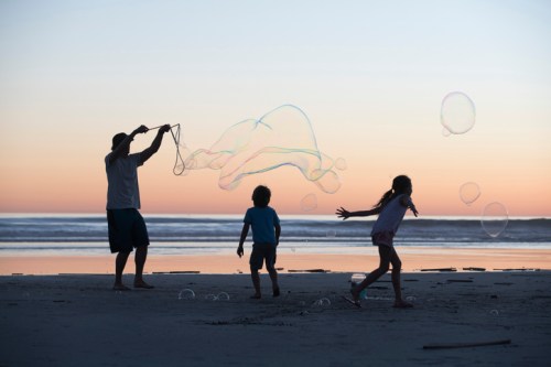 A silhouette of a person making bubbles while two children play on a beach at sunset.