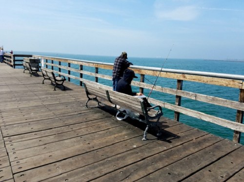Two people sit on a pier, one fishing and the other leaning back, with calm water and a clear sky in the background.