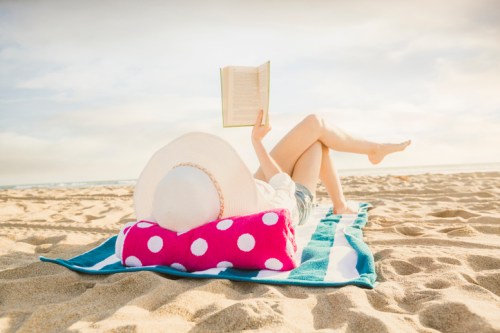 A person relaxes on a beach towel, reading a book while lying on the sand, wearing a large sun hat.