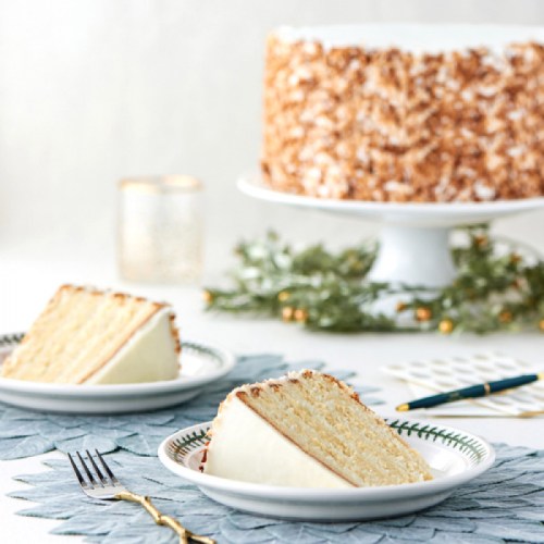 Two slices of cake on white plates, with a whole cake in the background, surrounded by festive greenery and decor.