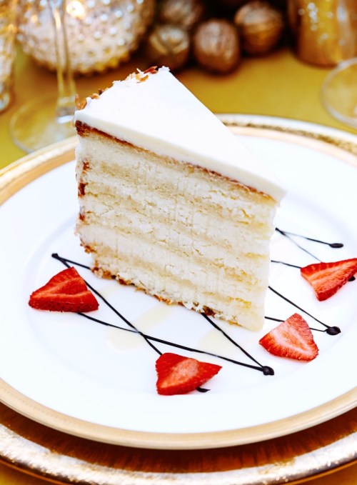 A slice of white cake with frosting, garnished with strawberries and chocolate drizzle, on a decorative plate.