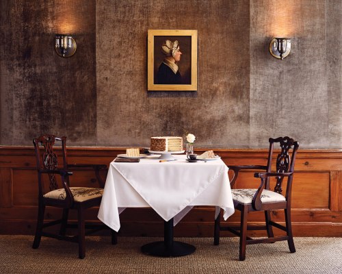 A cozy restaurant table set for two, featuring a white tablecloth, elegant chairs, and a framed portrait on the wall.