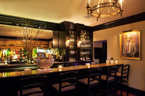 A stylish bar with a polished countertop, shelves of liquor, and a portrait on the wall, illuminated by warm lighting.