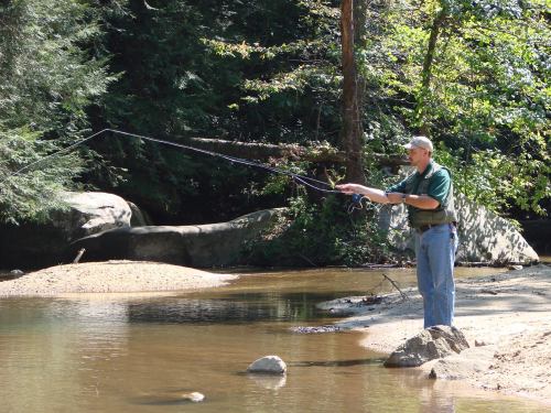 A person fishing by a calm river, surrounded by trees and rocks, enjoying a sunny day outdoors.