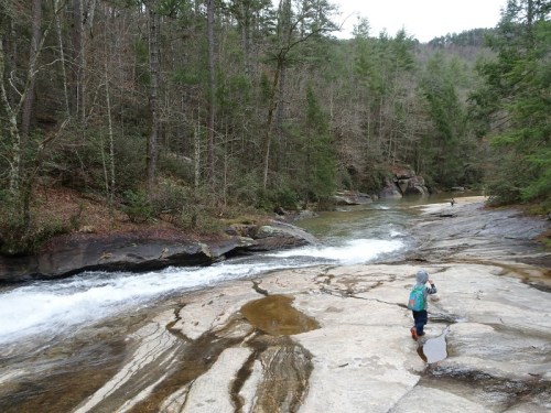A child stands on a rocky riverbank, surrounded by trees, with a flowing river in the background.