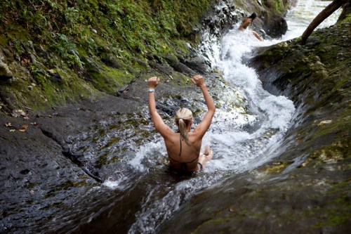 A person slides down a rocky water slide into a stream, surrounded by lush greenery. Another person is visible in the background.