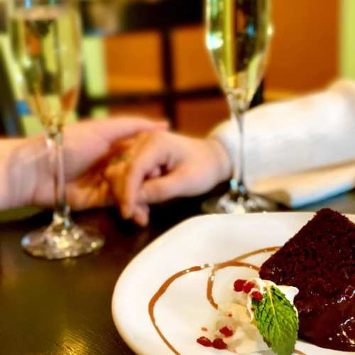 A close-up of two hands holding, with champagne flutes and a dessert plate featuring chocolate cake and cream.