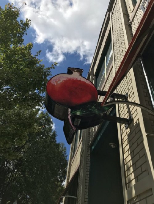 A colorful pomegranate sign hanging outside a building against a blue sky with clouds.