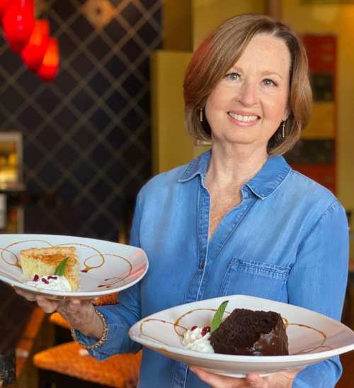 A woman smiles while holding two plates of dessert: a slice of pie and a chocolate cake with whipped cream.