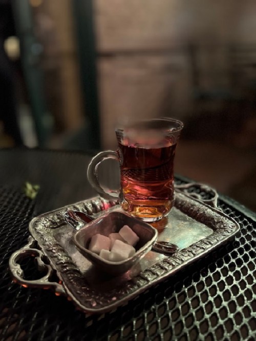 A glass of tea on a silver tray with a bowl of sugar cubes, set on a dark, textured surface.