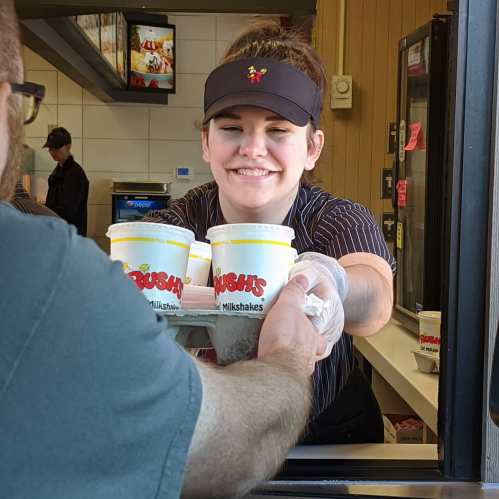 A smiling employee hands milkshake cups to a customer at a fast-food window.