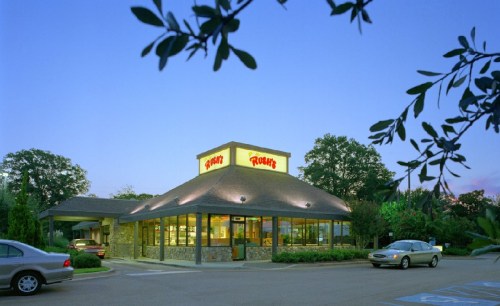 A fast-food restaurant with a stone exterior, illuminated at dusk, surrounded by trees and parked cars.