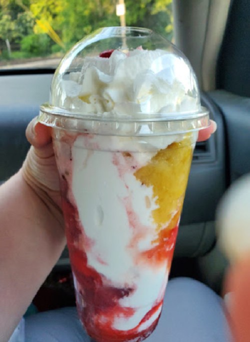 A hand holding a large dessert cup filled with layers of whipped cream, fruit, and colorful syrup.