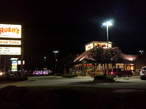 A brightly lit fast-food restaurant at night, featuring a drive-thru sign and parked cars.