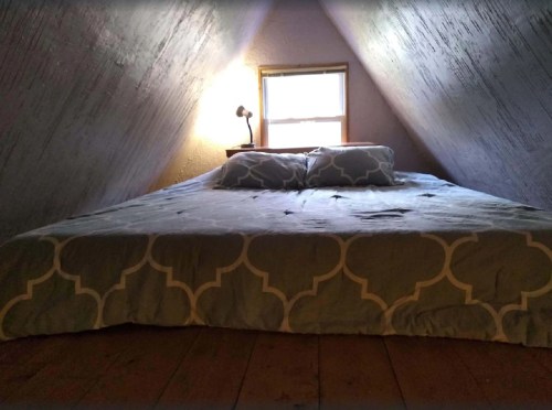 Cozy attic bedroom with a large bed, two pillows, and a lamp by a window, featuring sloped walls and wooden flooring.