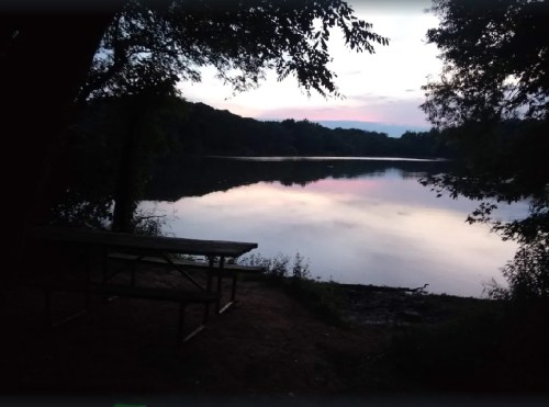 A serene lake at dusk, surrounded by trees, with a picnic table in the foreground and soft reflections on the water.
