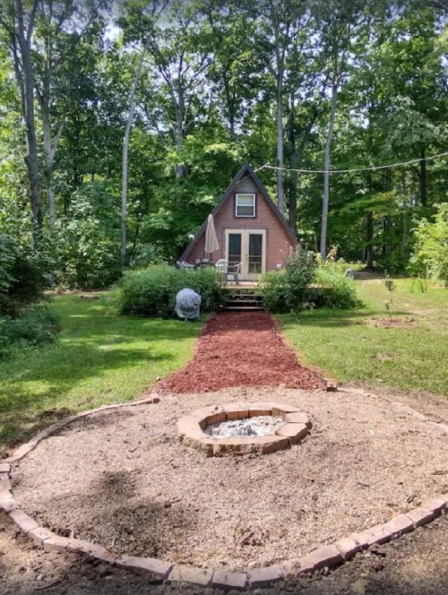A cozy A-frame cabin surrounded by trees, with a stone fire pit and a path leading to the entrance.