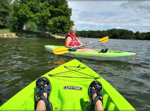 A person in a red life jacket kayaks on a lake, with a view of their feet in a green kayak in the foreground.
