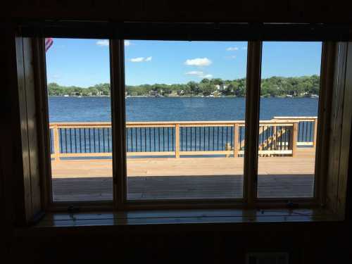 View of a serene lake through a large window, with a wooden deck and blue sky in the background.