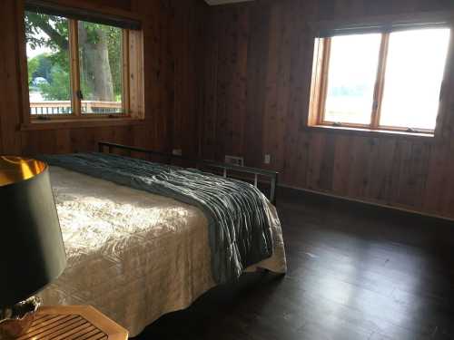 Cozy bedroom with wooden walls, a bed with a quilt, and large windows overlooking a lake.