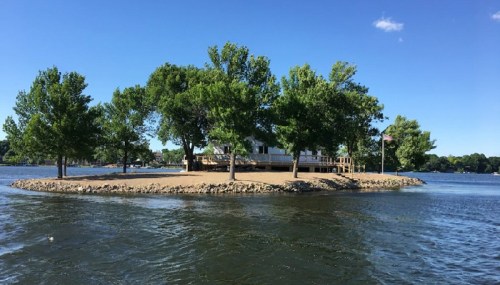 A small island with a house and trees surrounded by water under a clear blue sky.